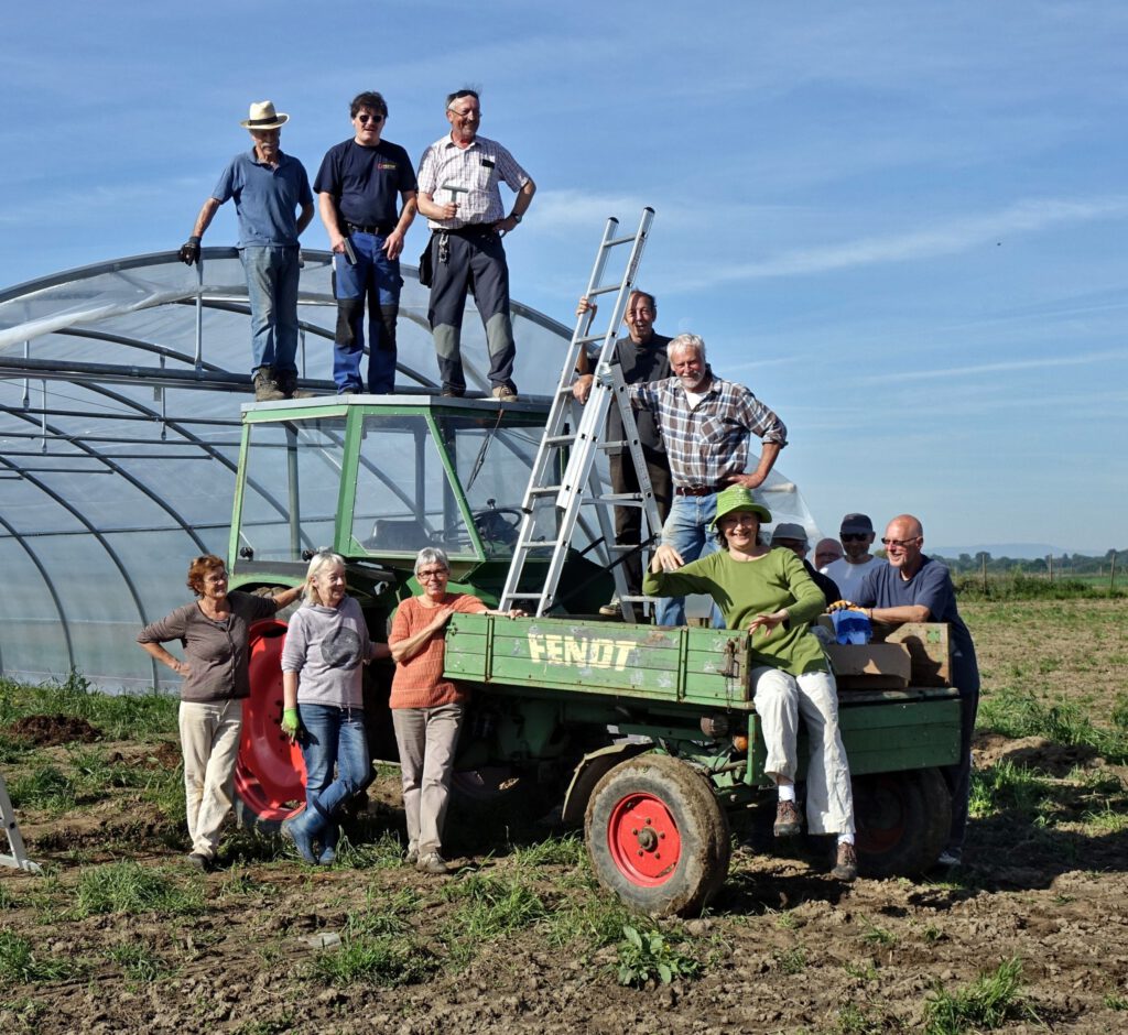 Eine Gruppe von Menschen posiert im Freien auf und um einen grünen Traktor auf einem Feld, mit einer Leiter und einem Gewächshaus im Hintergrund. Die Gruppe wirkt fröhlich und entspannt in einer ländlichen Umgebung.