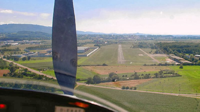 Blick aus dem Cockpit eines Kleinflugzeugs, das sich einer Landebahn nähert, wobei Felder, Bäume und Gebäude unter einem klaren blauen Himmel zu sehen sind. Der Propeller ist in der Mitte des Bildes unscharf.