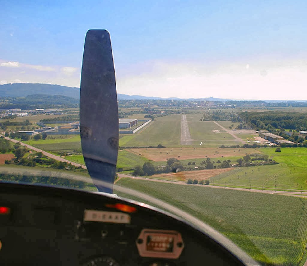 Blick aus dem Cockpit eines Kleinflugzeugs, das sich einer Landebahn nähert, wobei Felder, Bäume und Gebäude unter einem klaren blauen Himmel zu sehen sind. Der Propeller ist in der Mitte des Bildes unscharf.