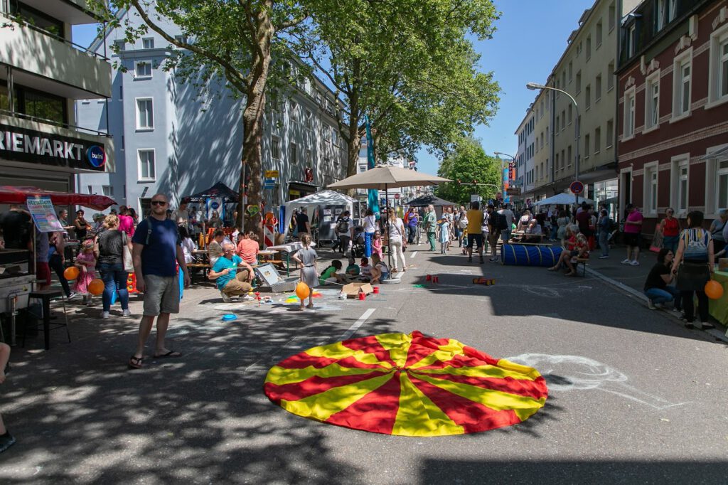 Ein lebendiges Straßenfest mit Menschen, die spazieren gehen, sich unterhalten und spielen. Bunte Dekorationen und Luftballons füllen die Szene. Ein großes rotes und gelbes Tuch liegt auf der Straße, und auf dem Bürgersteig sind Kreidezeichnungen zu sehen. Bäume spenden Schatten.