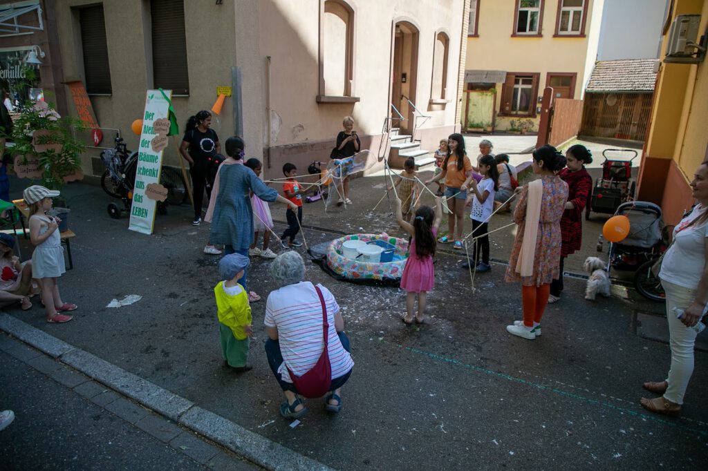 Kinder und Erwachsene versammeln sich um einen kleinen aufblasbaren Pool in einer Wohnstraße und spielen mit Schnüren und Wasser. Einige Erwachsene beaufsichtigen, während andere zuschauen oder sich unterhalten, und ein paar Kinder tragen unter dem sonnigen Himmel bunte Hüte.