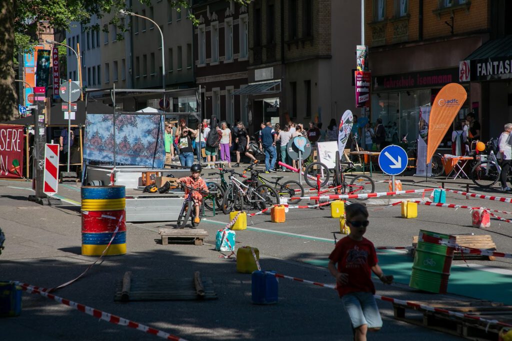 Kinder fahren Fahrrad und spielen auf einer für den Verkehr gesperrten Straße, auf der verschiedene Hindernisse, Fässer und Schilder aufgestellt sind. Menschen versammeln sich um Zelte und Stände auf dem Bürgersteig und genießen eine lebhafte Veranstaltung im Freien.