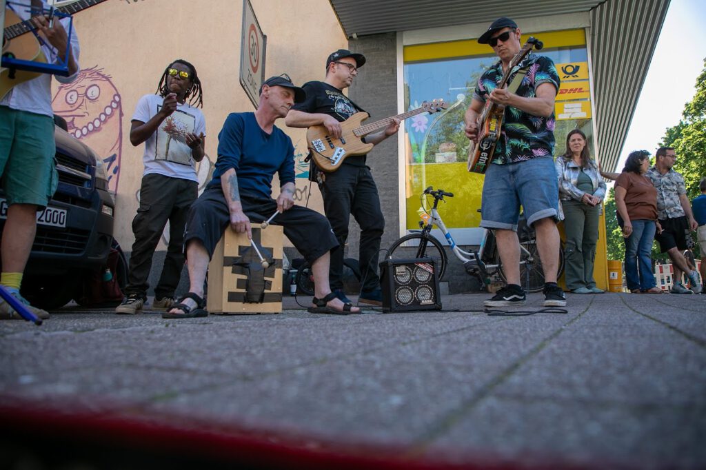 Eine Gruppe von Straßenmusikern spielt auf einem Bürgersteig mit Gitarren und einer Cajón-Trommel, während ein paar Leute vor einem Gebäude mit buntem Graffiti und einem gelben Fenster lächelnd zusehen.