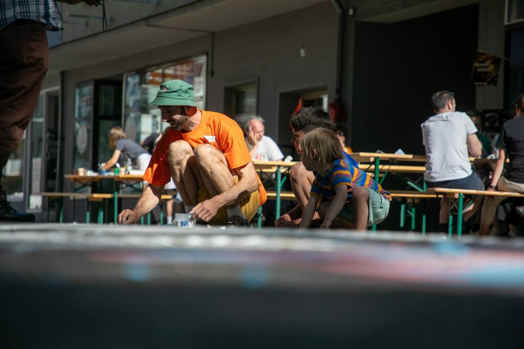 Ein Mann mit orangefarbenem Hemd und grünem Hut hockt neben einem kleinen Kind in gestreifter Kleidung und malt mit Kreide auf den Boden bei einer Veranstaltung im Freien, während im Hintergrund Menschen an Tischen sitzen.