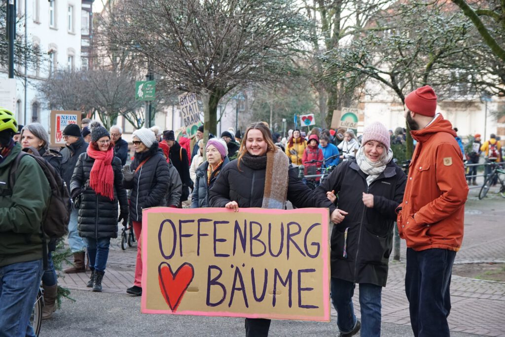 Eine Gruppe von Menschen hält bei einer Demonstration im Freien ein großes Schild mit der Aufschrift "Offenburg ♥ Bäume". Im Hintergrund sind Bäume und Gebäude zu sehen.