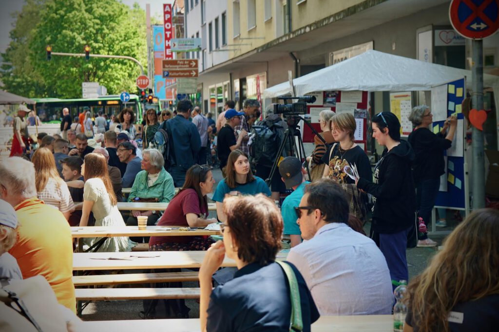Crowded street festival with people sitting at long wooden benches, tents and stalls lining the sidewalk, and a camera on a tripod in the foreground.
