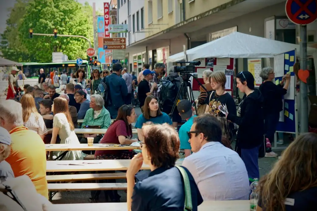 Crowded street festival with people sitting at long wooden benches, tents and stalls lining the sidewalk, and a camera on a tripod in the foreground.
