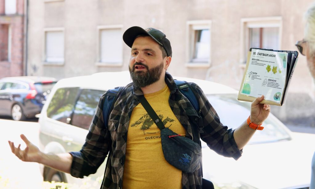 Bearded man wearing a cap and plaid shirt gestures with open arms while someone hands him a large brochure outdoors.