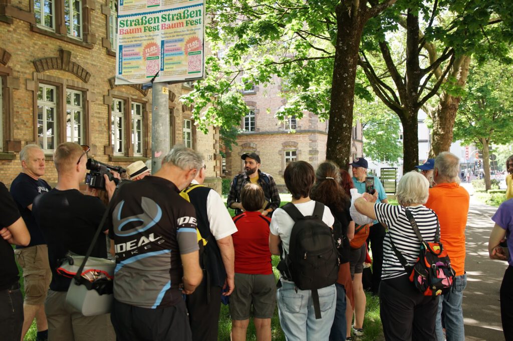 Eine Gruppe von Menschen hat sich draußen in der Nähe eines historischen Backsteingebäudes versammelt und hört einem Mann beim Reden zu. Mehrere tragen Rucksäcke, und eine Person filmt. Grüne Bäume spenden Schatten, und über der Gruppe hängen bunte Veranstaltungsplakate.