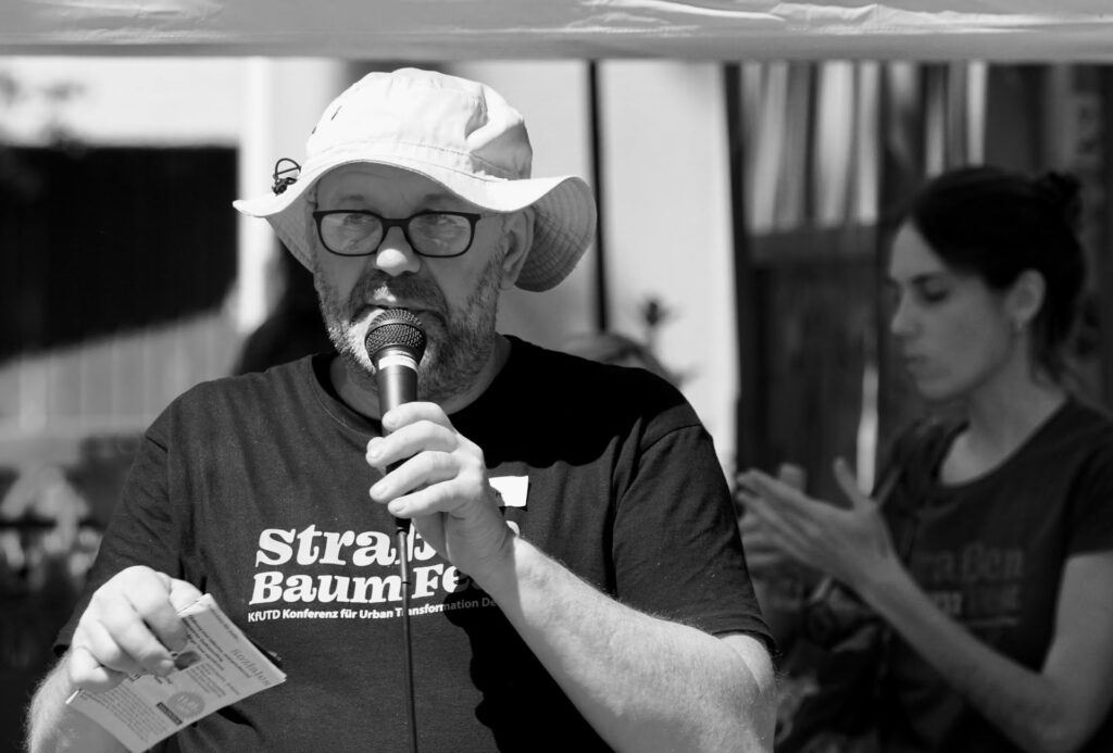 Man with a bucket hat and glasses speaks into a microphone, holding a flyer, outdoors; a person stands in the blurred background.