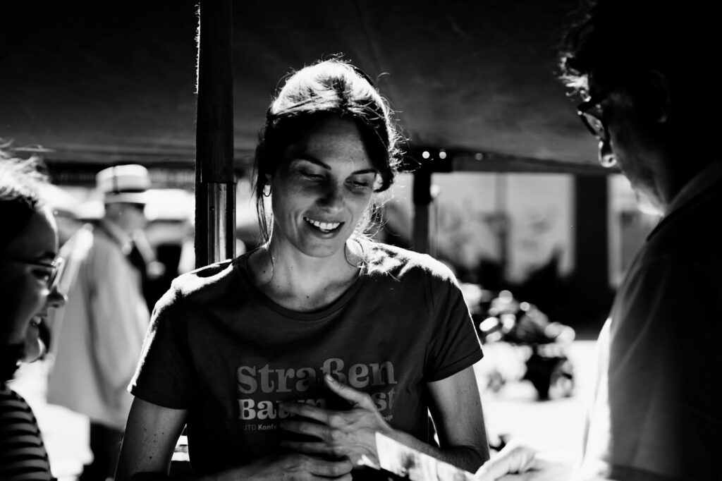 Smiling woman in a dark T-shirt stands outdoors, talking with people blurred in the background.