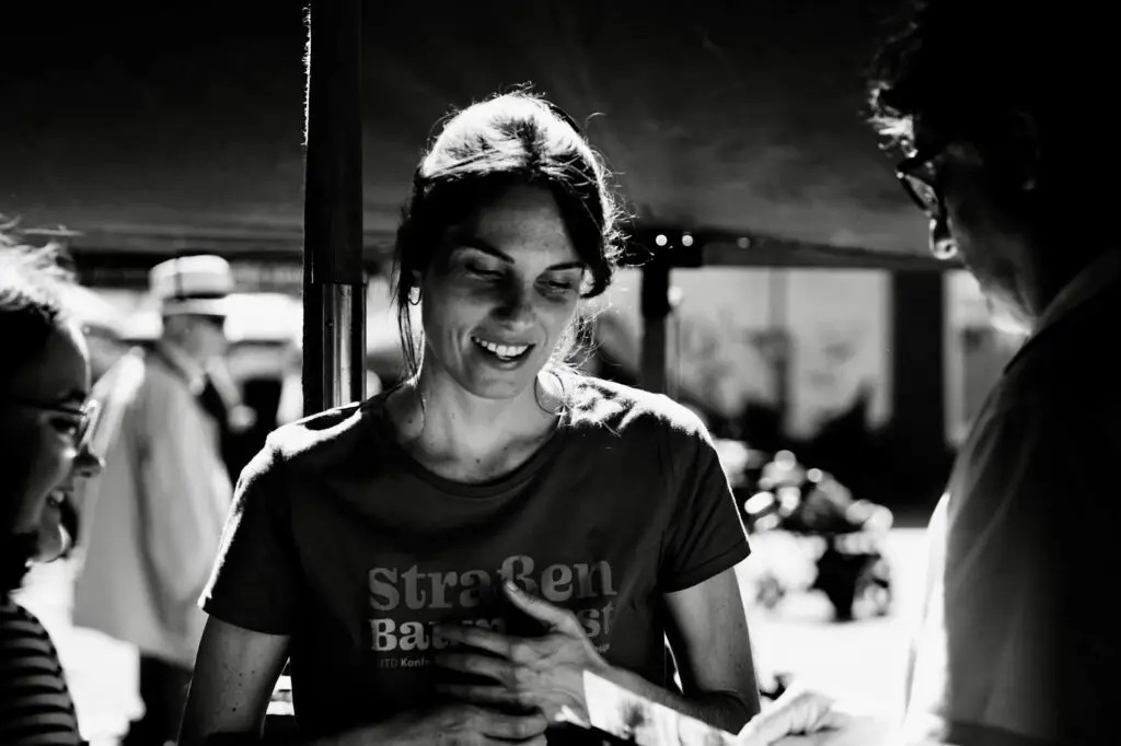 Smiling woman in a dark T-shirt stands outdoors, talking with people blurred in the background.