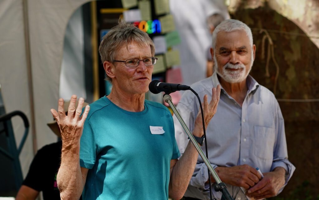 Two older men speak into a microphone at an outdoor event; the man in the teal shirt has a name tag reading 'Volker'.