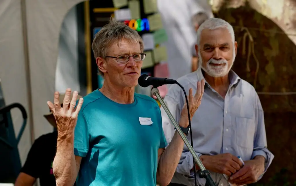 Two older men speak into a microphone at an outdoor event; the man in the teal shirt has a name tag reading 'Volker'.