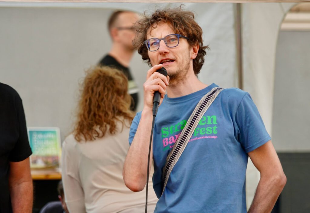 Man with blue glasses speaking into a microphone at an event, wearing a blue shirt and a striped bag strap across his chest.
