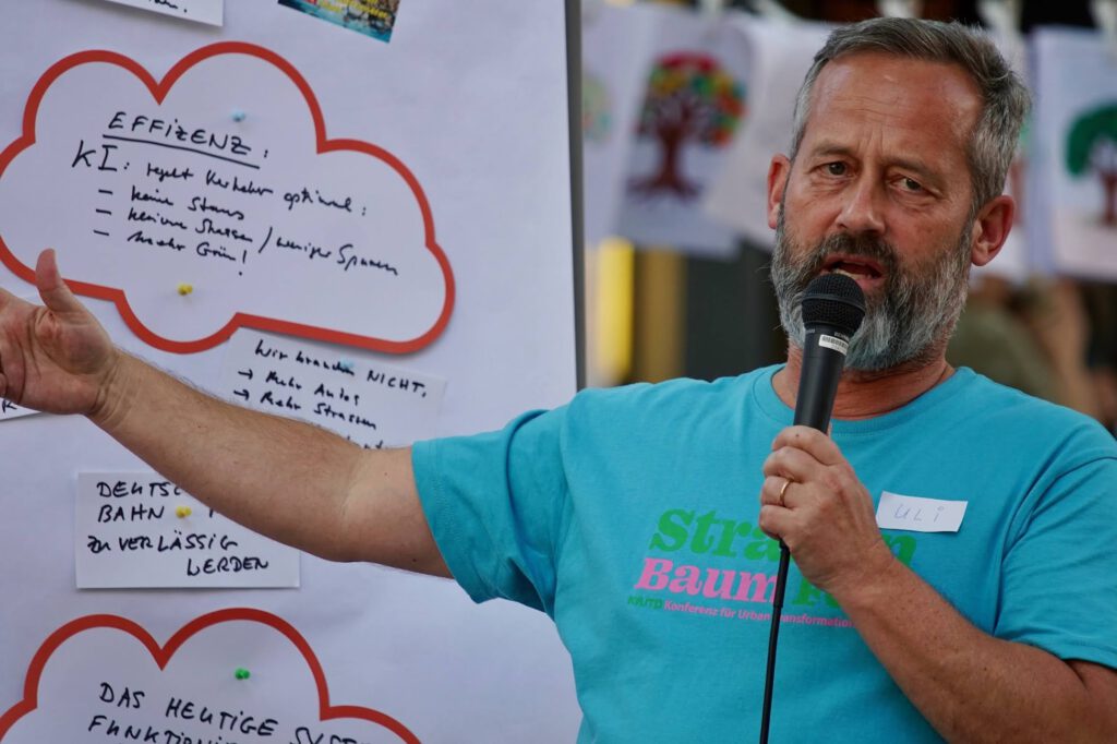 Man with gray beard speaks into a handheld microphone, standing beside a whiteboard filled with handwritten notes and red cloud outlines.