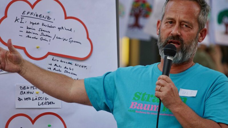 Uli Albicker. Ein Mann mit Bart, der ein türkisfarbenes Hemd trägt und ein Mikrofon in der Hand hält, gestikuliert, während er vor Plakaten mit handschriftlichen Notizen und Zeichnungen spricht. Er trägt ein Namensschild mit der Aufschrift Uli.