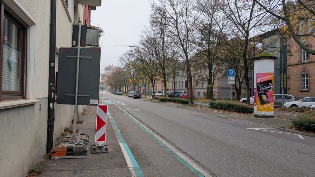 Eine ruhige, städtische Straße mit kahlen Bäumen im Herbst. Der Gehweg ist teilweise durch einen großen Stromkasten, ein Bauschild und Paletten blockiert. Entlang der Straße sind Autos geparkt, und auf beiden Seiten stehen Gebäude.