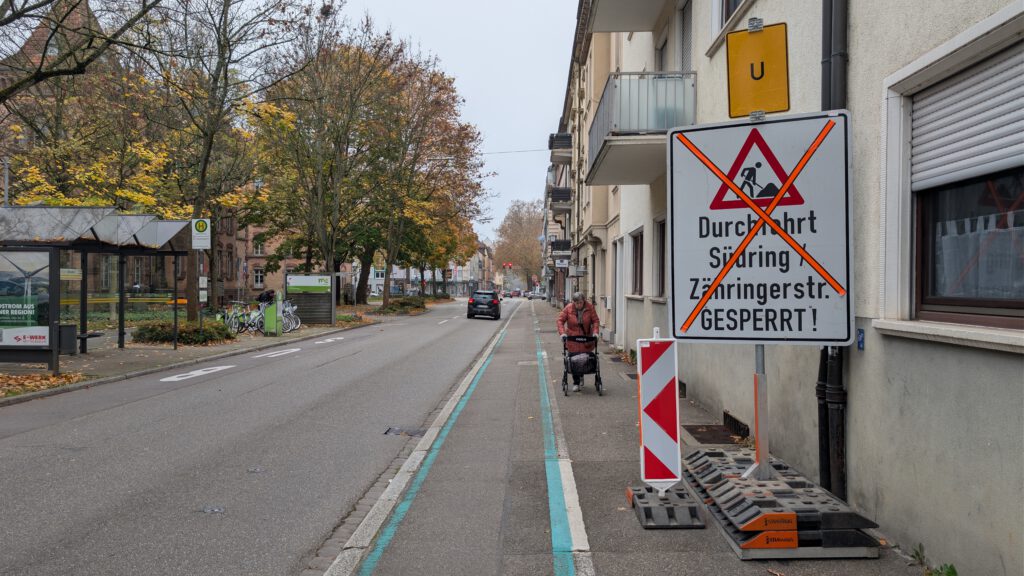 Eine Straße in einem Wohngebiet mit Gehweg und Radweg. Ein großes Schild zeigt an, dass die Straße gesperrt ist (rot durchgestrichen). Eine Person im Rollstuhl fährt auf dem Gehweg. Bäume mit Herbstlaub säumen die Straße.