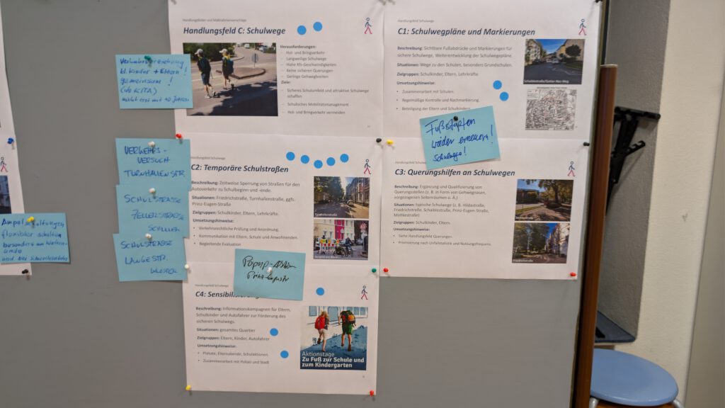 An einer Pinnwand hängen mehrere gedruckte Dokumente über Schulwege, die mit blauen Haftnotizen und Fotos von Kindern, die zu Fuß unterwegs sind, versehen sind. Die Papiere sind mit bunten Stecknadeln befestigt.