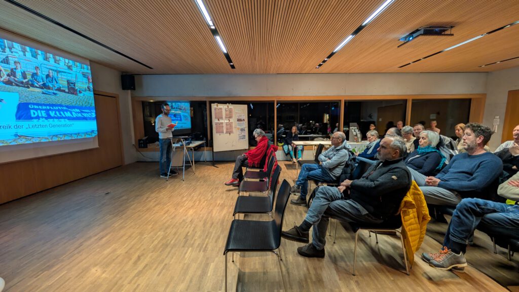 Eine Gruppe von Personen sitzt in einem hell erleuchteten Raum und sieht zu, wie eine Person einen Vortrag hält, dessen Folien auf eine große Leinwand projiziert werden. Einige Teilnehmer sind engagiert, andere sitzen still da.