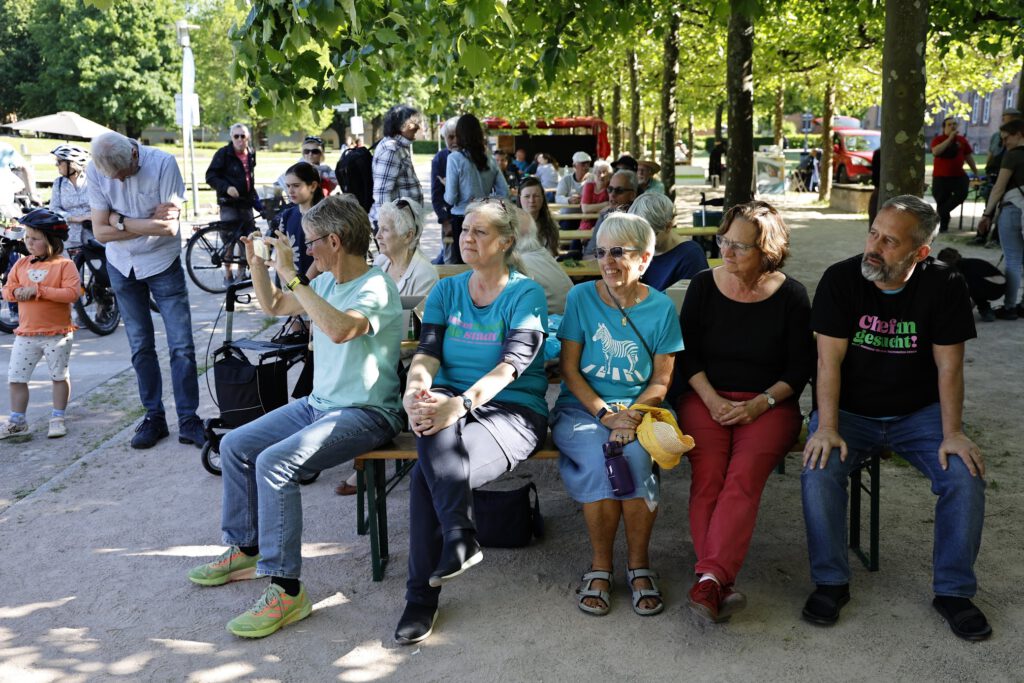 Eine Gruppe von Menschen, meist ältere Erwachsene, sitzt auf Bänken im Freien unter Bäumen und beobachtet etwas aus dem Off. Andere stehen oder gehen in der Nähe.