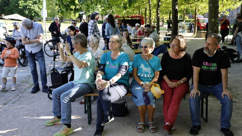 Eine Gruppe von Menschen, meist ältere Erwachsene, sitzt auf Bänken im Freien unter Bäumen und beobachtet etwas aus dem Off. Andere stehen oder gehen in der Nähe.
