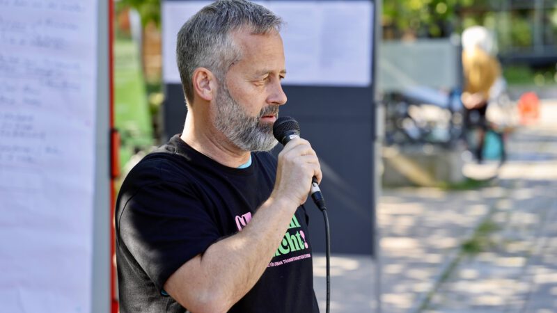 Ein Mann mit kurzen grauen Haaren und einem Bart spricht im Freien in ein Mikrofon und steht neben einer Schautafel mit Plakaten. Er trägt ein schwarzes T-Shirt, und das Sonnenlicht fällt durch die grünen Bäume im Hintergrund.
