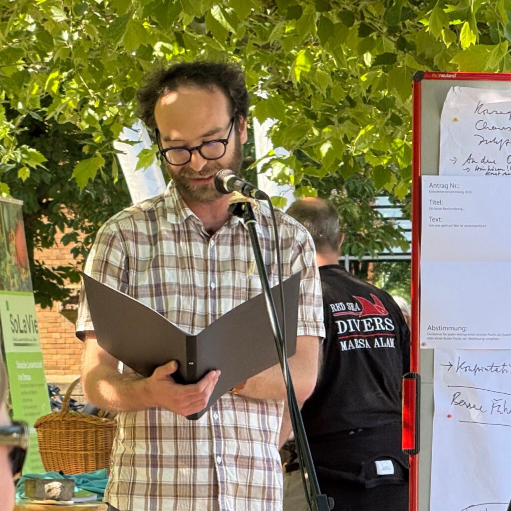 Mathias Gnädinger mit Brille und kariertem Hemd steht im Freien an einem Mikrofon und liest aus einem schwarzen Ordner vor. Hinter ihm sind Menschen, Bäume und eine Tafel mit handschriftlichen Notizen zu sehen.