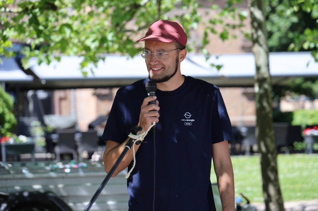 Man wearing a red cap and glasses speaks into a handheld microphone outdoors, with trees and blurred seating in the background.