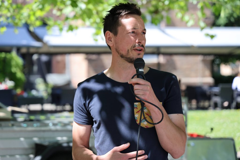 Man in a navy shirt speaks into a handheld microphone outdoors with trees in the background.