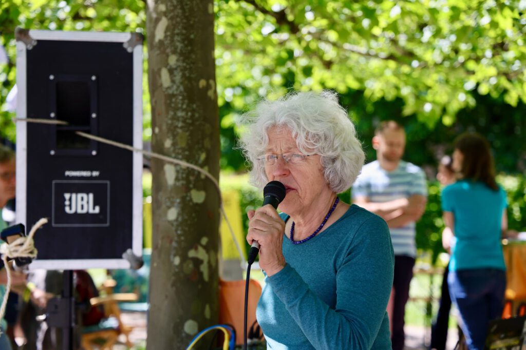 Older woman with white hair singing into a microphone at an outdoor event, with a JBL speaker visible in the background.