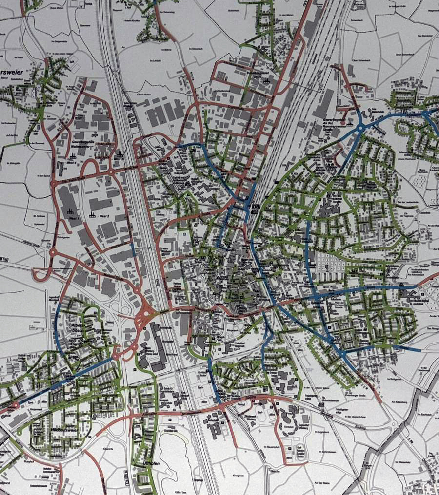 Ein detaillierter Stadtplan, der Straßen, Eisenbahnlinien und verschiedene Gebäudeblöcke in verschiedenen Farben zeigt, mit grünen Flächen für Parks, blauen Linien möglicherweise für Flüsse oder Routen und roten Linien, die Hauptstraßen markieren. Es ist kein Text deutlich lesbar.