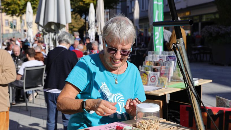 Eine ältere Frau mit kurzen grauen Haaren und Sonnenbrille lächelt, während sie an einem Tisch im Freien sitzt und Holzmünzen aus einem Glas sortiert. Im Hintergrund sind Menschen bei einer Veranstaltung im Freien an einem sonnigen Tag versammelt.