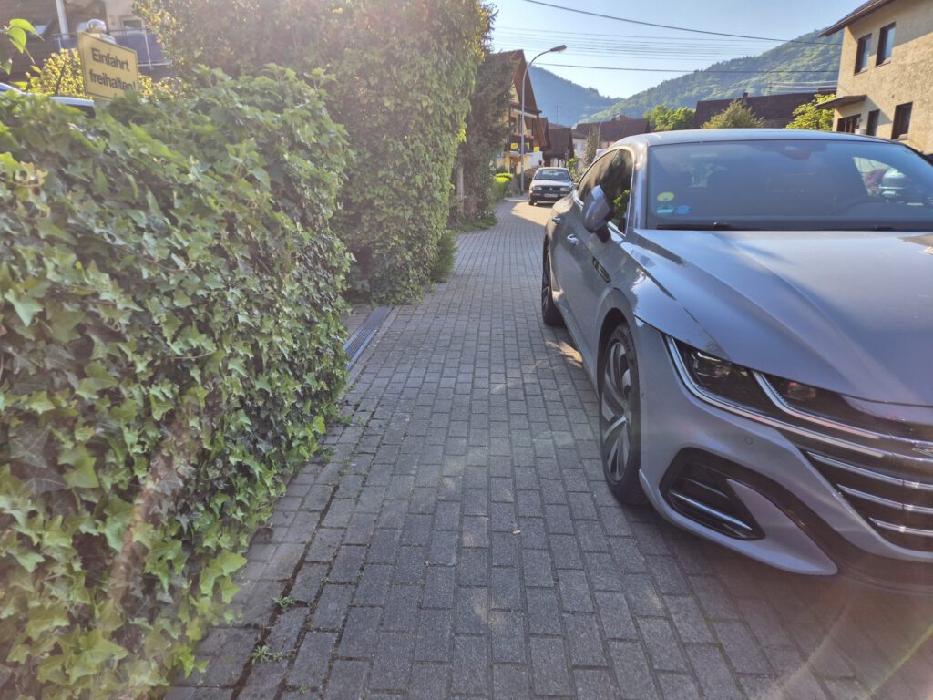 Ein graues Auto ist an einem sonnigen Tag in einem Wohnviertel mit Hügeln im Hintergrund teilweise auf einem schmalen Ziegelstein-Gehweg neben einer Hecke geparkt.