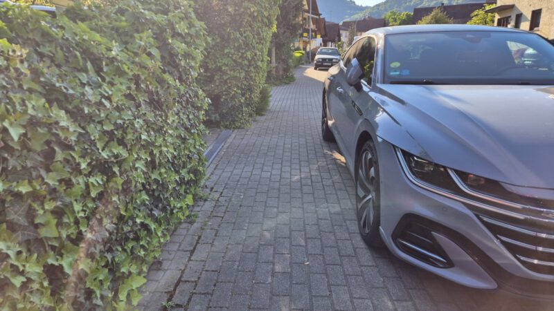 Ein graues Auto ist an einem sonnigen Tag in einem Wohnviertel mit Hügeln im Hintergrund teilweise auf einem schmalen Ziegelstein-Gehweg neben einer Hecke geparkt.