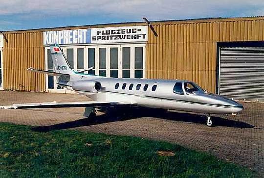 Ein kleiner silberner Jet mit dem Kennzeichen D-ICIT parkt auf einer gepflasterten Fläche vor einem hellbraunen Gebäude mit großen Fenstern und dem Schild KONPRECHT FLUGZEUG-SPRITZWERFT.