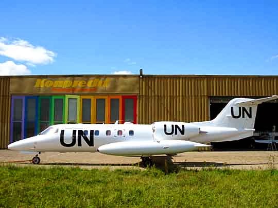 Ein kleiner weißer Jet mit großen schwarzen UN-Buchstaben auf dem Rumpf und dem Heck steht auf dem Gras vor einem braunen Hangar mit regenbogenfarbenen Paneelen und dem Zeichen Kompact.