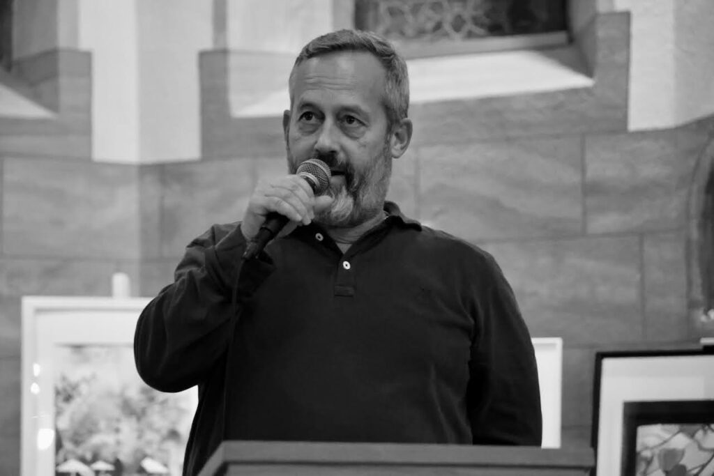 Uli Albicker spricht in ein Mikrofon, während er in einem Haus steht. Er trägt ein dunkles langärmeliges Hemd. Im Hintergrund sind gerahmte Bilder und strukturierte Steinwände zu sehen. Das Foto ist in schwarz-weiß gehalten.