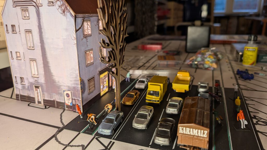 Eine Miniaturstadtszene mit Spielzeugautos, Lastwagen und Figuren in der Nähe eines Papiermodellgebäudes. Die Fahrzeuge sind entlang der Straße geparkt, und mehrere kleine Figuren gehen oder stehen auf dem Bürgersteig. Die Gebäudewand hat bemalte Fenster.