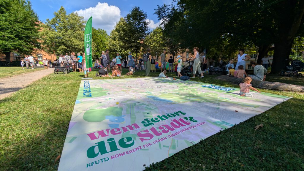 Ein großer Stadtplan mit der Aufschrift "Wem gehört die Stadt?" liegt auf einer Wiese in einem Park. Menschen, darunter auch Kinder, versammeln sich um sie herum, einige sitzend oder stehend, mit Bäumen und Gebäuden im Hintergrund an einem sonnigen Tag.