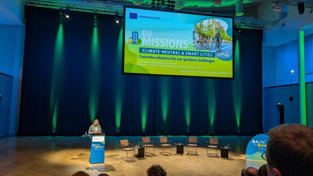 Eine Rednerin steht an einem Podium auf einer Bühne mit sieben leeren Stühlen und grüner Beleuchtung. Hinter ihr zeigt ein großer Bildschirm die EU-Missionen: Klimaneutrale und intelligente Städte" auf einer Veranstaltung der Europäischen Union.
