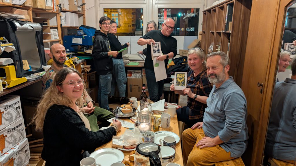 Eine Gruppe von Menschen sitzt und steht um einen Holztisch in einem gemütlichen, unordentlichen Raum und teilt Essen und Getränke. Einige lächeln in die Kamera, andere sehen sich Abzüge an; im Hintergrund stehen Bücher und Zubehör in den Regalen.