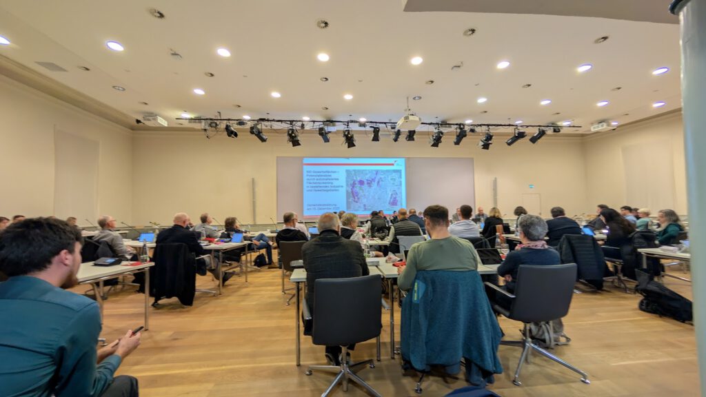 Eine Gruppe von Personen sitzt an Tischen vor einem großen Bildschirm in einem Konferenzraum und nimmt an einer Präsentation oder einem Workshop teil. Der Raum hat Holzböden, hohe Decken und eine moderne Beleuchtung.
