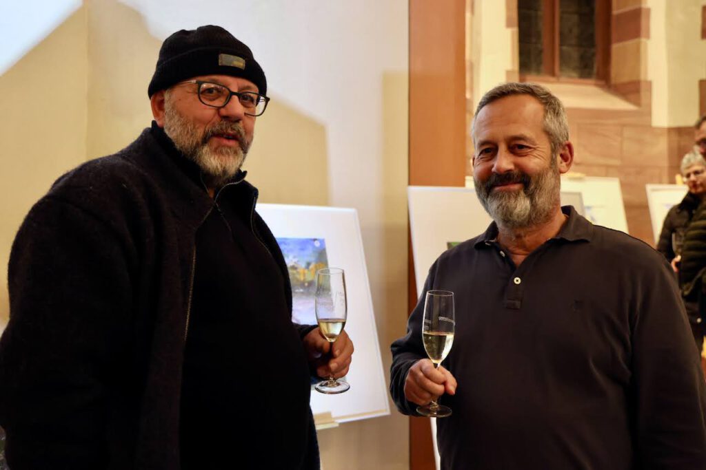 Zwei Männer stehen in einem Haus, halten Gläser mit Champagner in der Hand und lächeln in die Kamera. Der eine trägt eine schwarze Mütze, eine Brille und eine dunkle Jacke, der andere hat einen Bart und trägt ein dunkles Poloshirt. Im Hintergrund sind Kunstwerke ausgestellt.