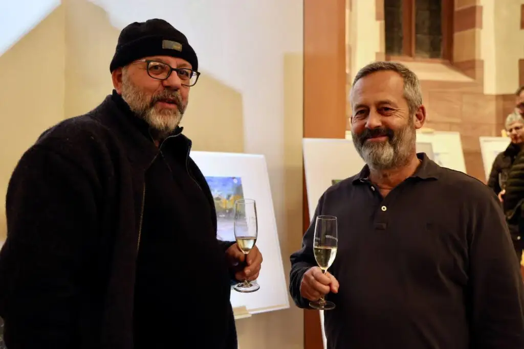 Zwei Männer stehen in einem Haus, halten Gläser mit Champagner in der Hand und lächeln in die Kamera. Der eine trägt eine schwarze Mütze, eine Brille und eine dunkle Jacke, der andere hat einen Bart und trägt ein dunkles Poloshirt. Im Hintergrund sind Kunstwerke ausgestellt.