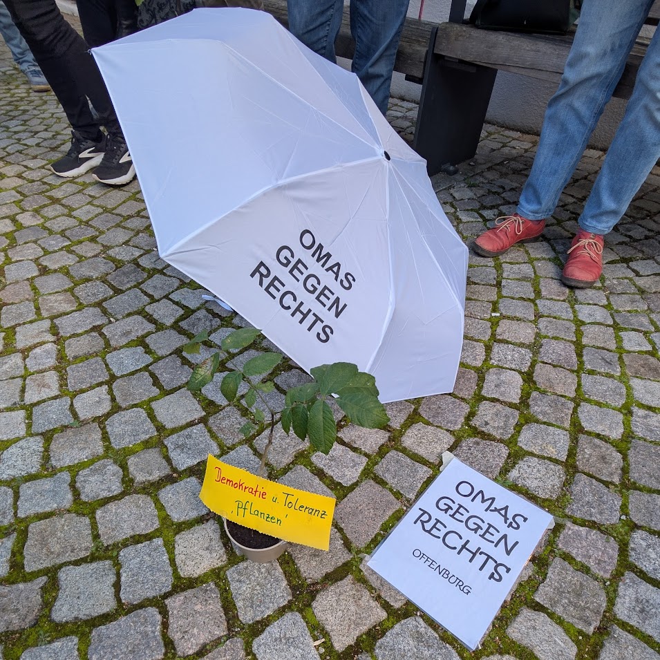 Ein weißer Regenschirm mit der Aufschrift OMAS GEGEN RECHTS beschattet eine Pflanze auf Pflastersteinen. In der Nähe stehen ein Schild mit der Aufschrift Demokratie u. Toleranz - pflanzen und ein Flugblatt mit demselben Slogan. Drei Personen stehen in der Nähe.