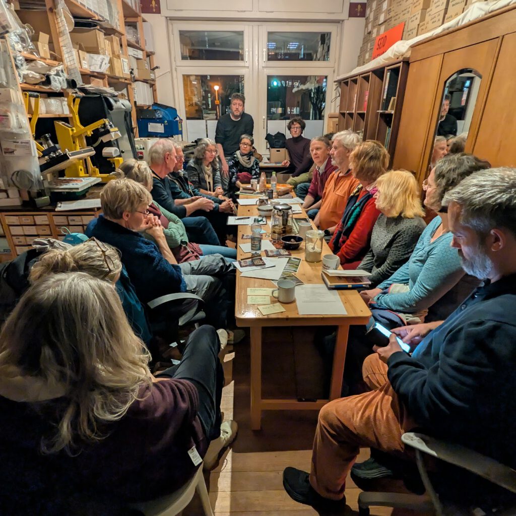 Eine Gruppe von Menschen sitzt um einen langen Tisch in einem gemütlichen, unordentlichen Raum und unterhält sich. An den Wänden stehen Regale mit Kisten und Papieren, und einige Leute halten Papiere in der Hand oder benutzen Telefone. Warme Innenraumbeleuchtung.