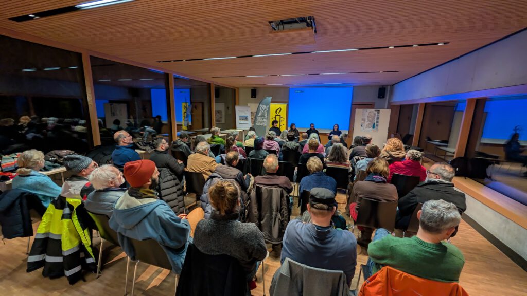 In einem modernen, holzgetäfelten Raum mit großen Fenstern und blauer Beleuchtung sitzt eine Gruppe von Menschen in Reihen vor einer Reihe von Rednern. Einige der Anwesenden tragen Winterkleidung und Mützen.