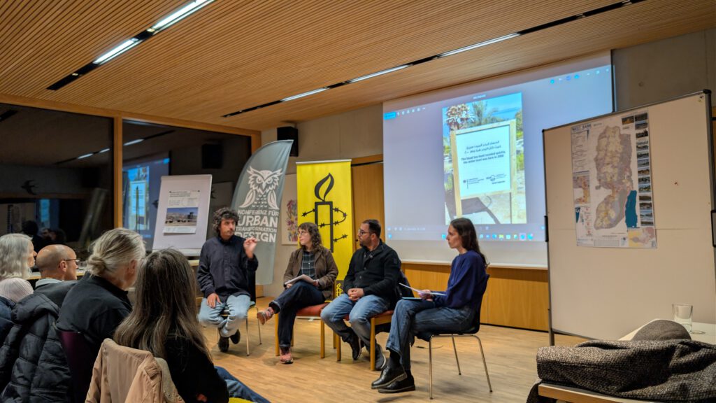 Vier Personen sitzen auf einem Bühnenpult vor einem Publikum, hinter ihnen wird auf einem großen Bildschirm eine Präsentation gezeigt. Zwei Banner, eines mit einer Eule und eines mit dem Logo von Amnesty International, stehen im Hintergrund.